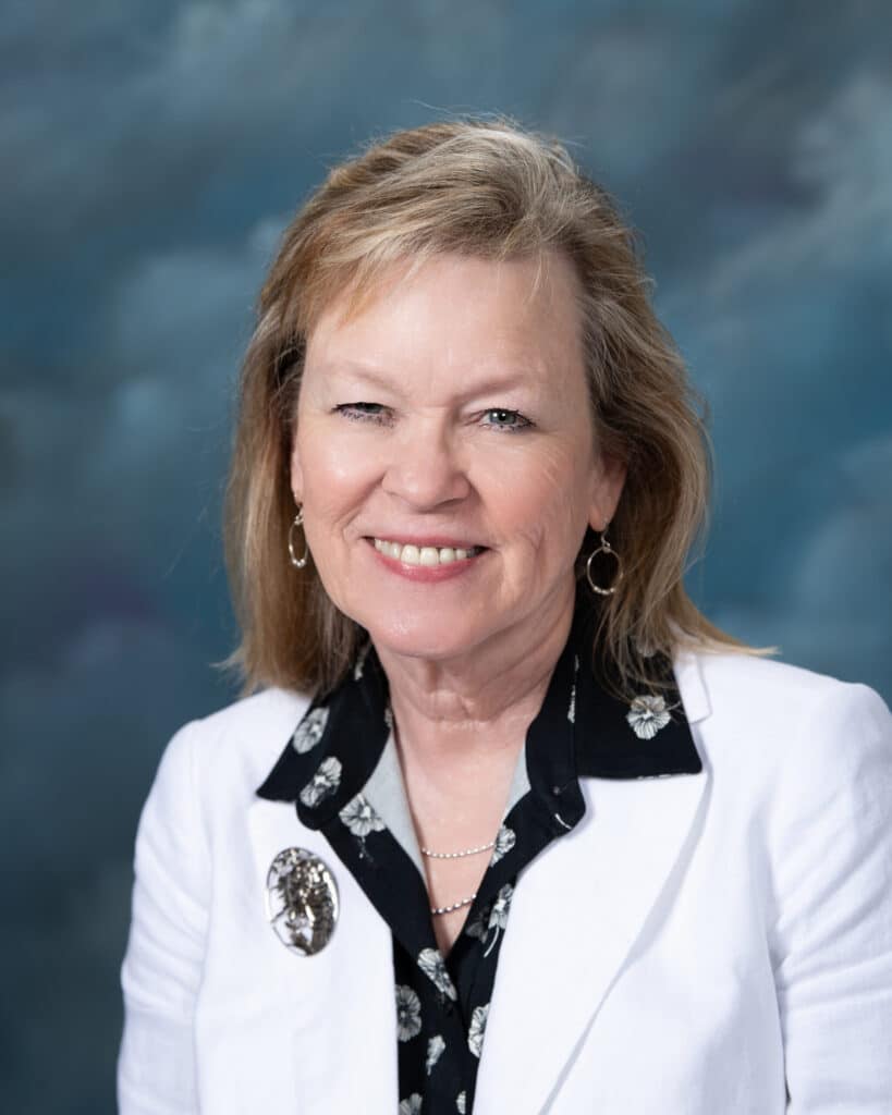 A smiling woman with shoulder-length blonde hair is wearing a white blazer over a black patterned blouse. She has hoop earrings and a brooch. The background is a soft, blue and gray gradient.