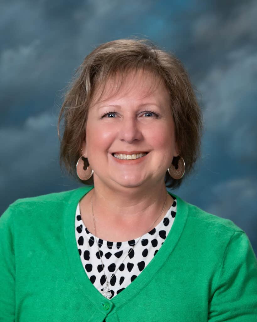 A woman with short light brown hair, wearing a green cardigan over a black-and-white polka dot top, smiles at the camera against a blue and gray cloudy background.