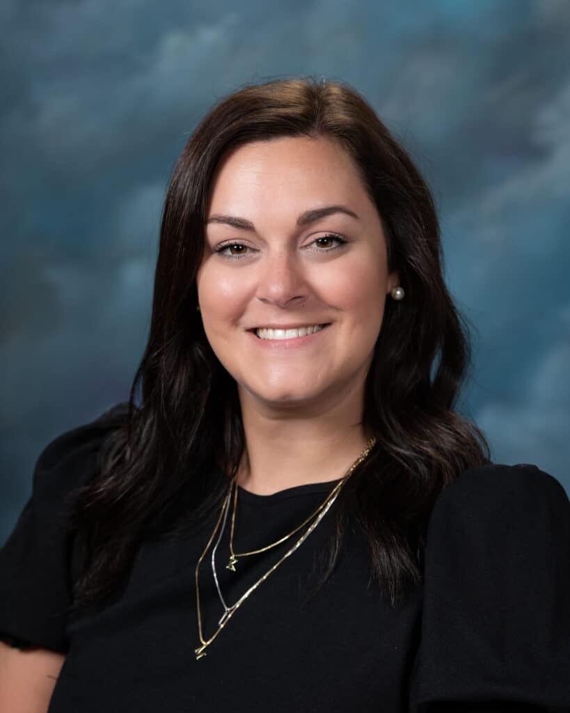 A woman with long dark brown hair and light skin, wearing a black top and gold layered necklaces, smiles in front of a blue and gray background.