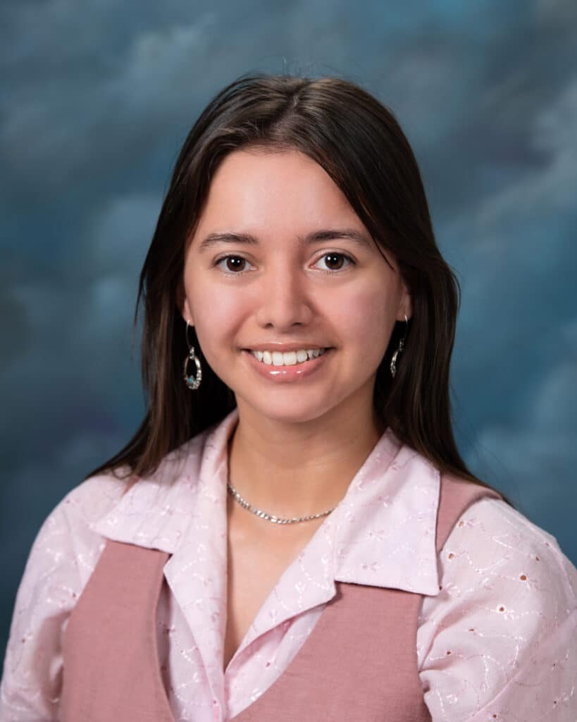 A young woman with long dark hair, wearing a light pink blouse and vest, smiles at the camera against a softly blurred blue and gray background.