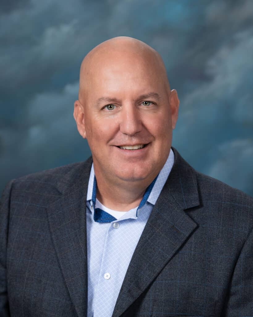A bald man wearing a blue blazer and light blue shirt smiles at the camera, posed against a cloudy blue and gray studio background.