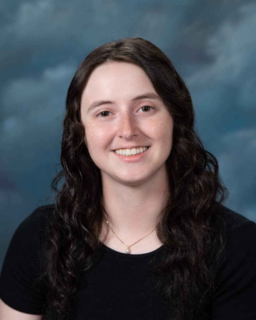 A young woman with long, wavy brown hair smiles at the camera. She is wearing a black top and a delicate necklace. The background is a softly blurred blue and gray studio backdrop.