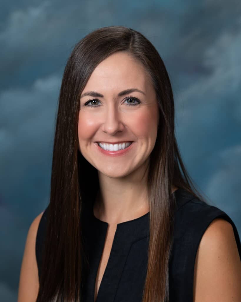 A woman with long straight brown hair, wearing a sleeveless black top, smiles at the camera in front of a cloudy blue background.