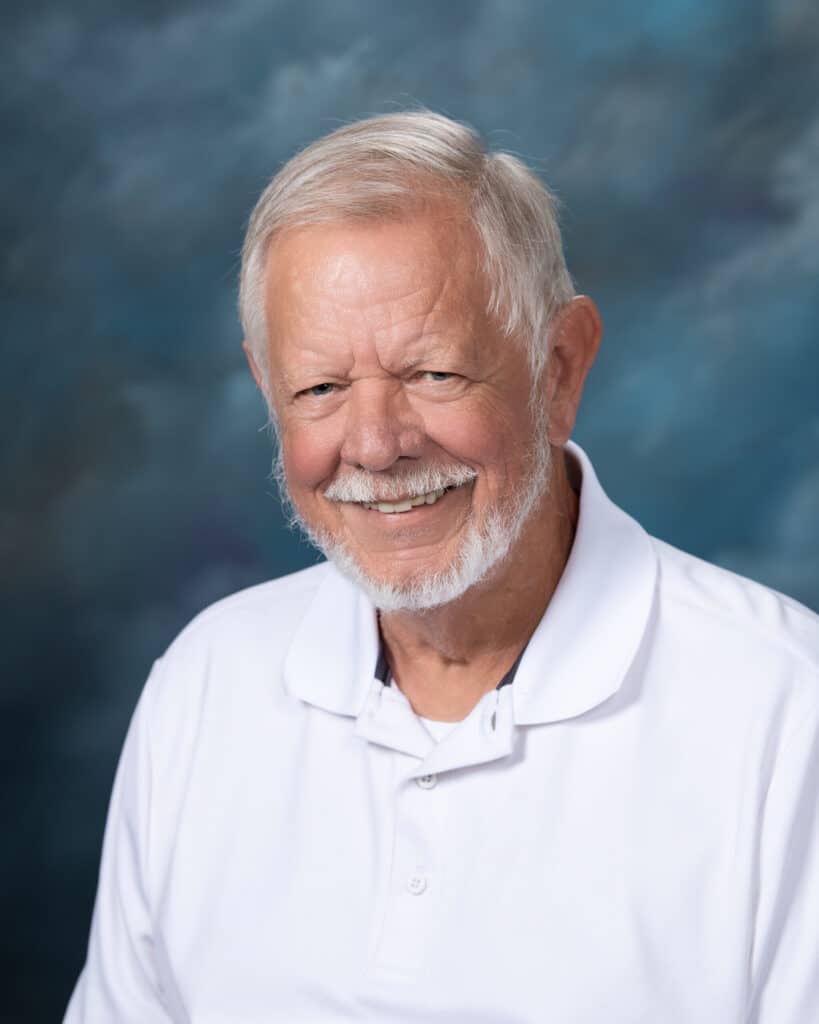 An older man with white hair and a beard smiles warmly. He is wearing a white polo shirt and is posed in front of a blue and gray mottled studio backdrop.