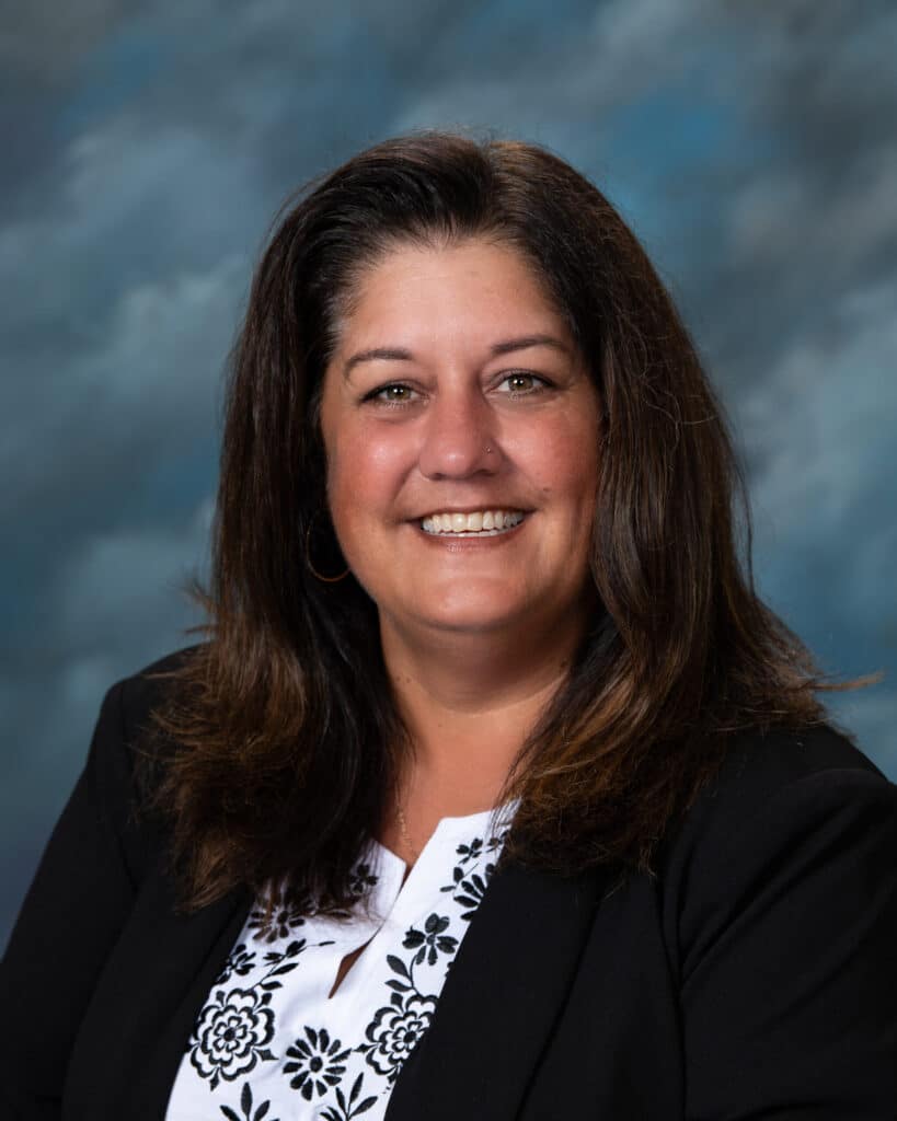 A woman with long brown hair wearing a black blazer over a white blouse with black floral designs smiles at the camera in front of a blue and gray mottled background.