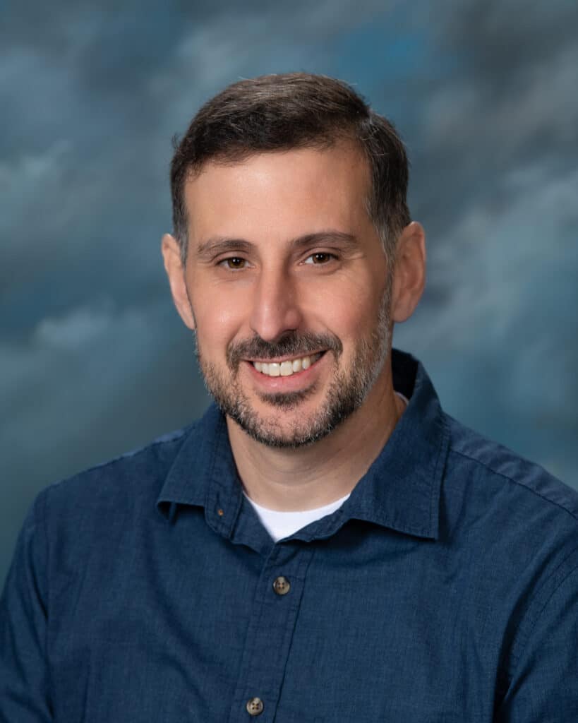 A man with short dark hair and a beard, wearing a blue button-up shirt over a white t-shirt, smiles in front of a cloudy blue and gray studio backdrop.