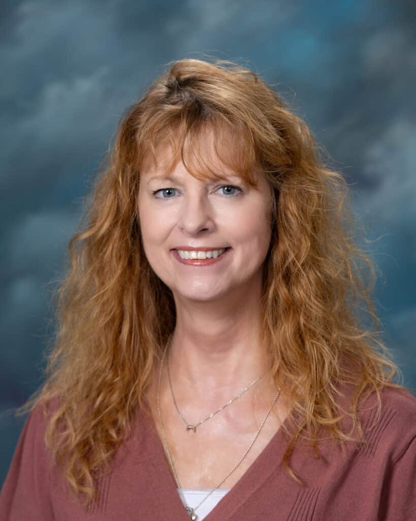 A woman with long, wavy, reddish-blonde hair smiles at the camera. She is wearing a mauve top and layered necklaces, with a cloudy blue and gray backdrop behind her.