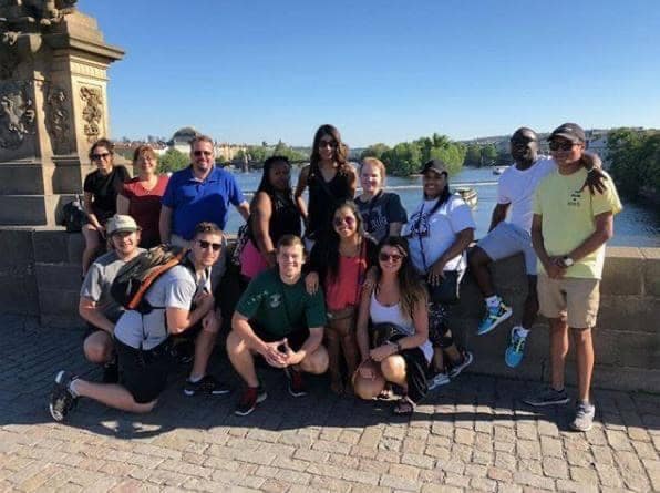 Group of students posing on a bridge with boats and a river in the background