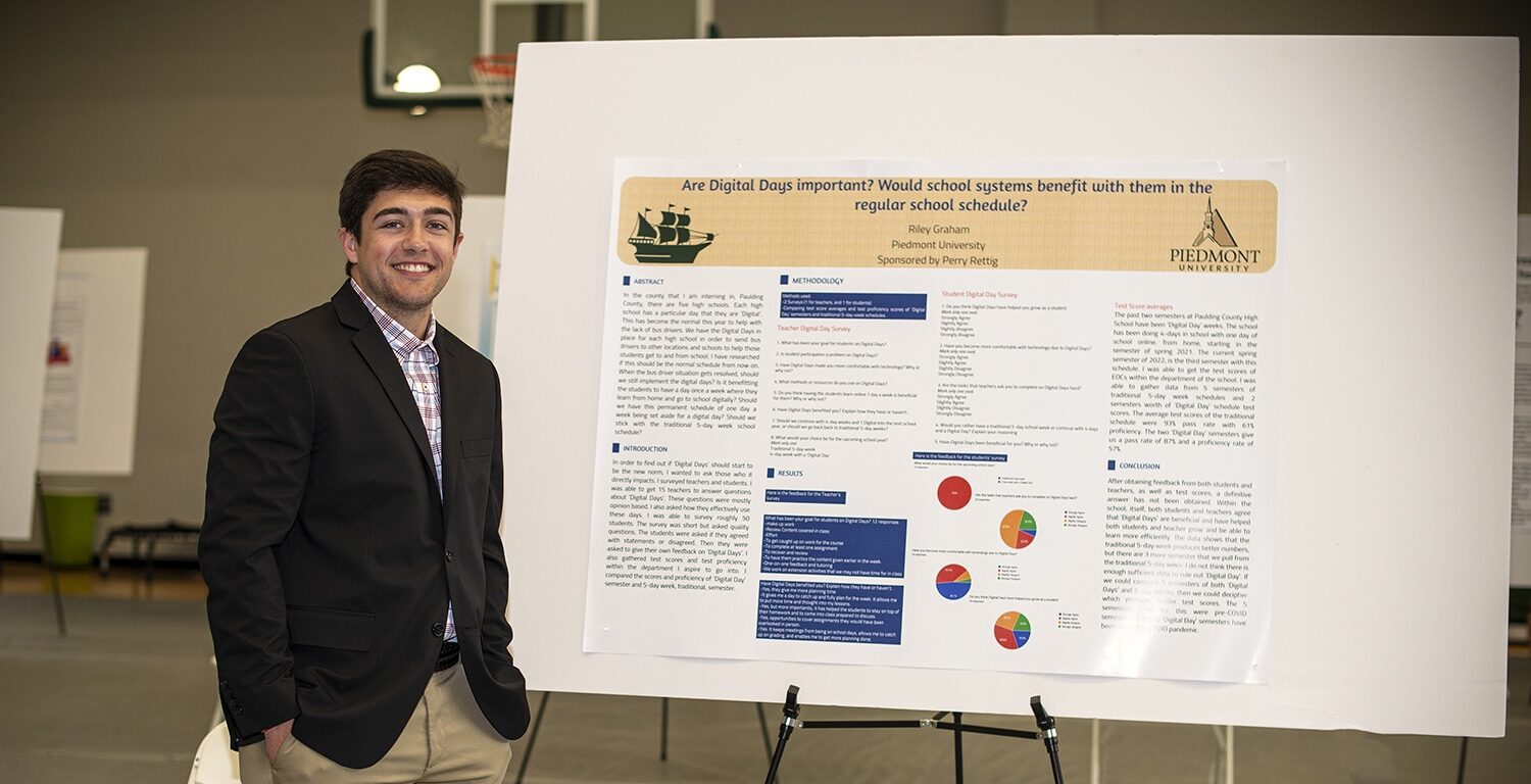 A young man in a black blazer and khaki pants stands smiling next to a large presentation board titled “Are Digital Days Important? Would school systems benefit…”. The board displays text, charts, and the Piedmont University logo.